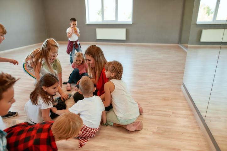 Full length portrait of positive dance teacher talking to group of little girls and boys sitting on the floor in studio. Relationship between teacher and kids. Choreography class. Dance school