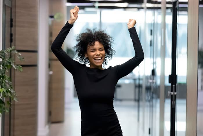 Overjoyed young African American female employee or worker dance in office hallway enjoy Friday fun. Happy smiling millennial biracial businesswoman celebrate job success or work achievement.